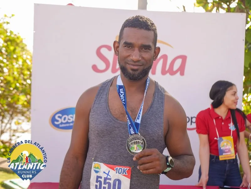 Un corredor masculino con una medalla de un 10K en la Carrera del Atlántico del Atlantic Runners Club, con un fondo que incluye un cartel de la carrera y un árbol.