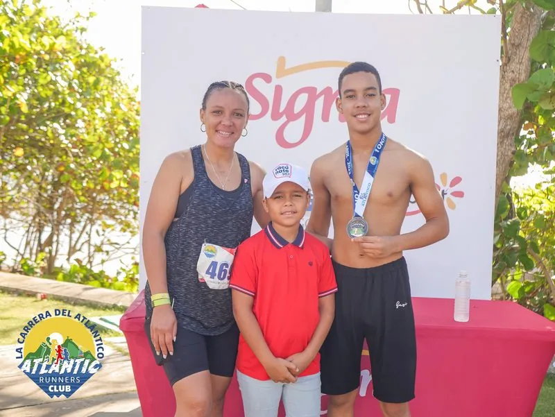 Una familia compuesta por un adulto y dos niños posa con medallas y bibs de carrera en un evento atlético. El fondo muestra un cartel con el nombre del evento y el logo del club.