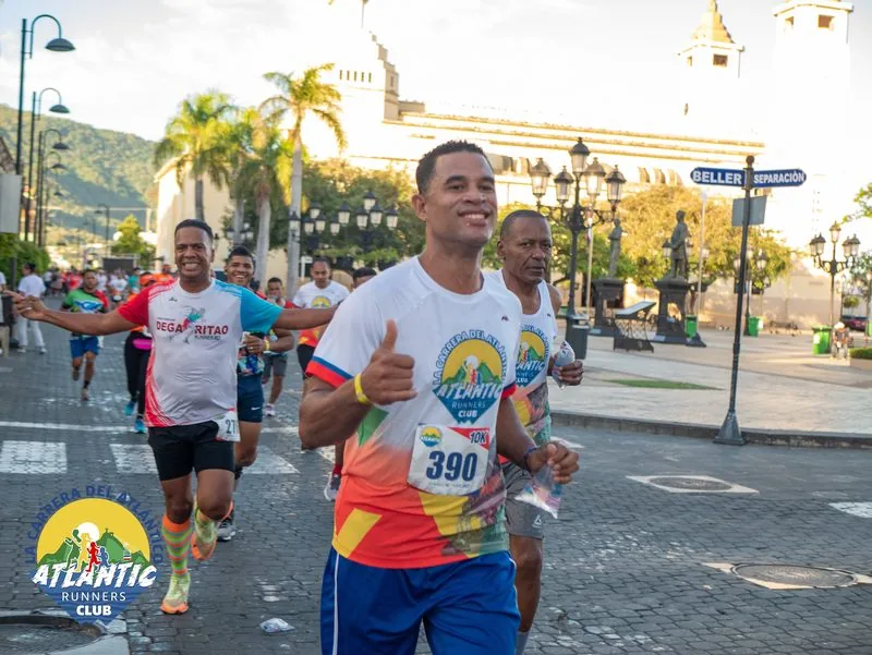 Un corredor con un número de participación en el pecho corre en una carrera de atletismo en una plaza con edificios históricos al fondo.