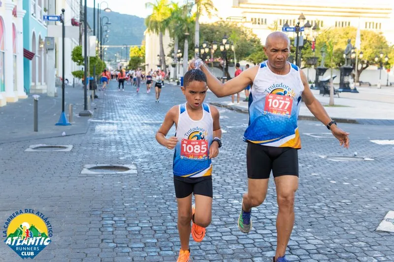 Una imagen de un padre y su hijo corriendo juntos en una carrera atlética en la República Dominicana. El niño lleva el número 1085 y el hombre el número 1077.
