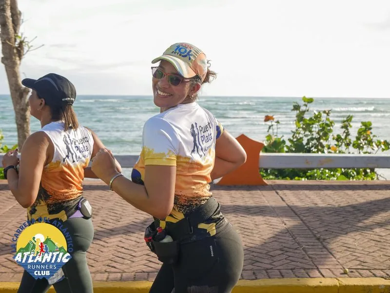 Damas con camisetas de atletismo y gorras, posando cerca del océano.