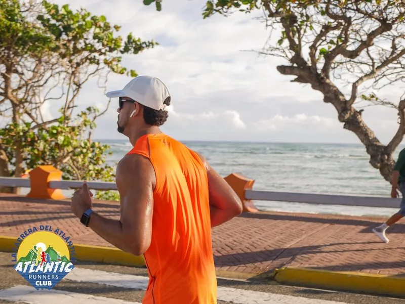 Un corredor masculino con un atuendo deportivo naranja y un sombrero blanco, corriendo en un entorno costero con el mar en el fondo.