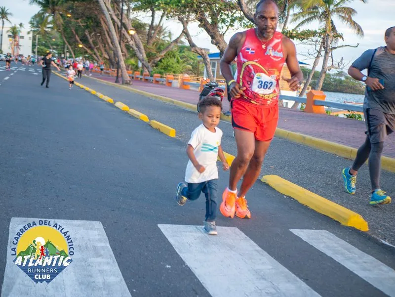 Una imagen de un corredor adulto y un niño cruzando una línea de meta en una carrera atlética. El corredor adulto lleva un número de participación y el niño camina junto a él.