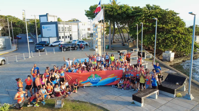 Vista aérea del Atlantic Runners Club reunido en el malecón de Puerto Plata, sosteniendo la bandera del club frente al mar.