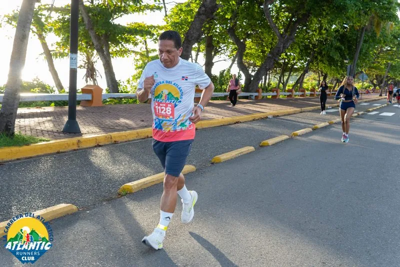 Un corredor con un número de participación en la camiseta corre en una carrera en un entorno verde con árboles y calles adoquinadas.