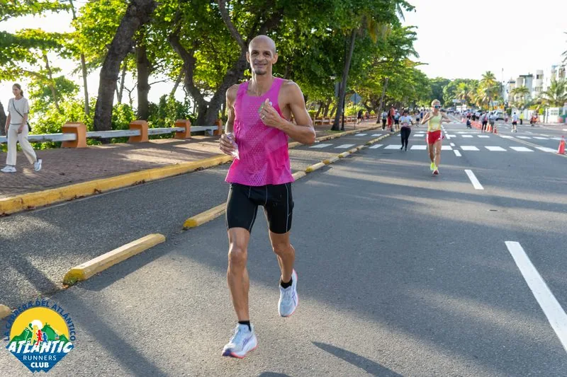 Un corredor masculino en un atuendo deportivo rosado corre con energía en una calle arbolada. Otros corredores se ven en el fondo.